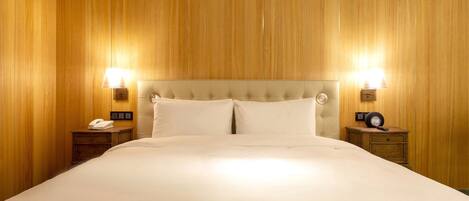 Habitación de hotel con una cama grande, paneles de madera en las paredes, dos mesitas de noche con lámparas y un reloj sobre una de ellas.