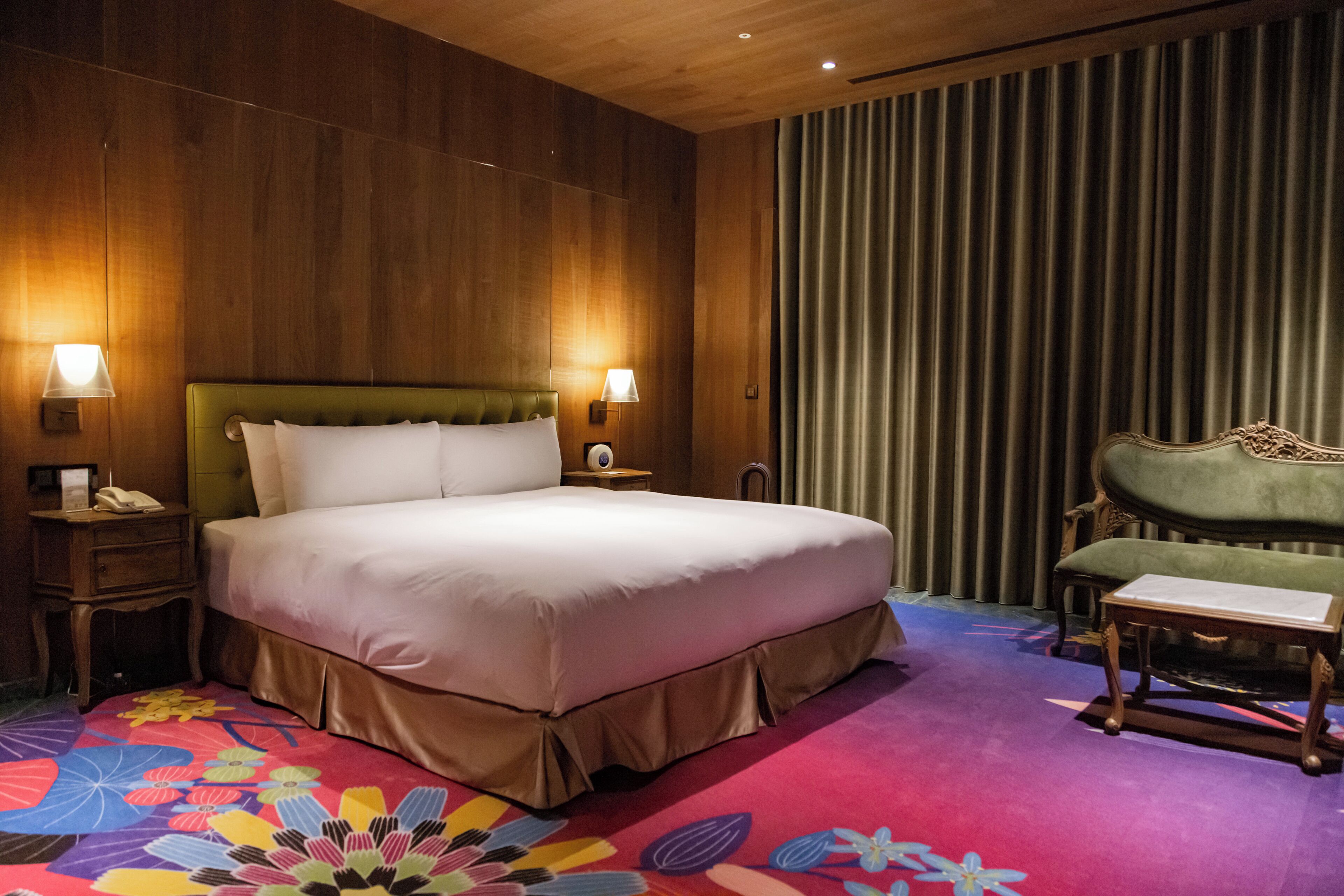 A hotel room with a large bed, two bedside lamps, a green armchair, a small table, and a colorful carpet.