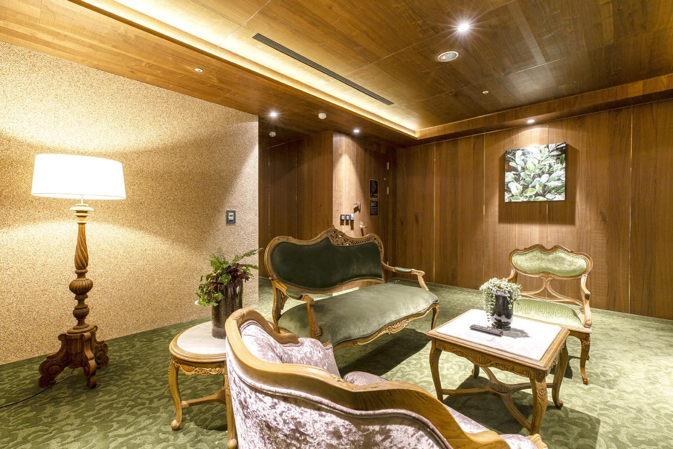 A room with a wooden ceiling, green upholstered chairs, a table with a vase, and a lamp.