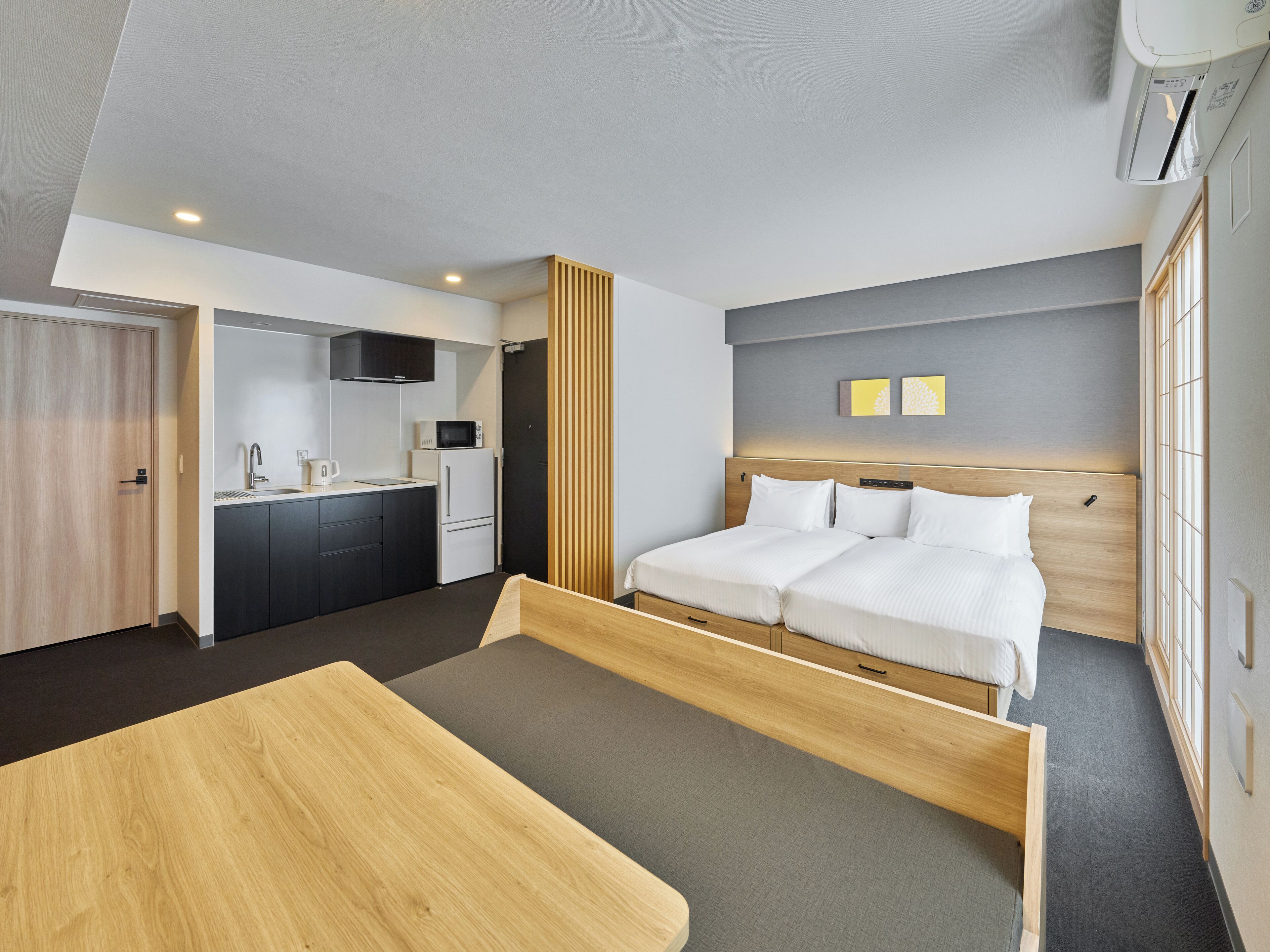 A modern hotel room with a bed, a wooden desk, a kitchenette, and a large window.