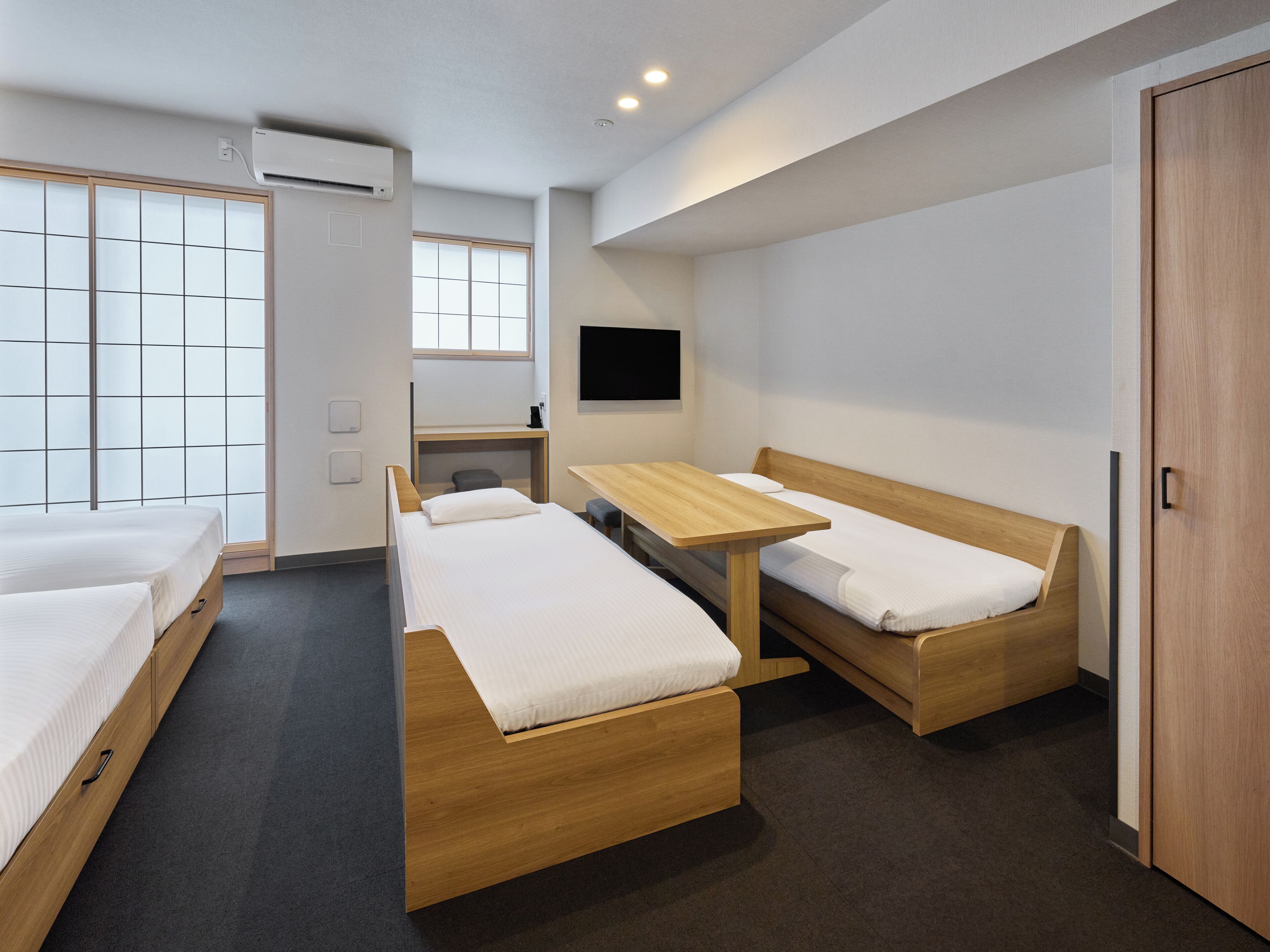 A modern hotel room with two single beds, a wooden desk, a chair, a TV mounted on the wall, and a sliding door leading to a balcony.