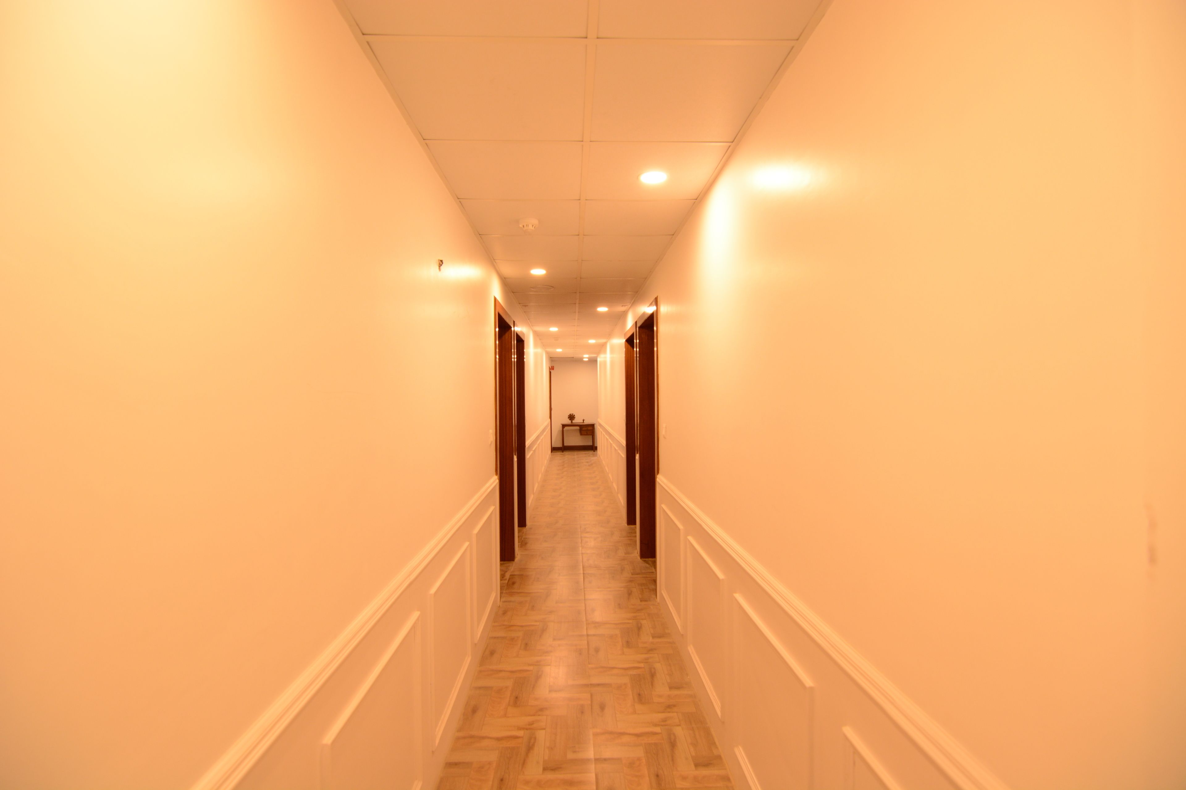 A long, narrow hallway with wooden flooring and white walls, leading to a room at the end.