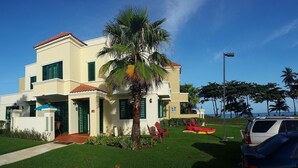 A two-story building with a palm tree in the front yard, a red chair, and a kayak. - VillaNicky Rincon, PR 2,800 Sqft Beach Front Oasis  (Rincon)