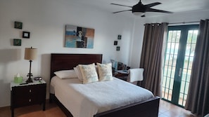 A bedroom with a bed, a nightstand, a lamp, a ceiling fan, a window with curtains, and artwork on the wall. - VillaNicky Rincon, PR 2,800 Sqft Beach Front Oasis  (Rincon)