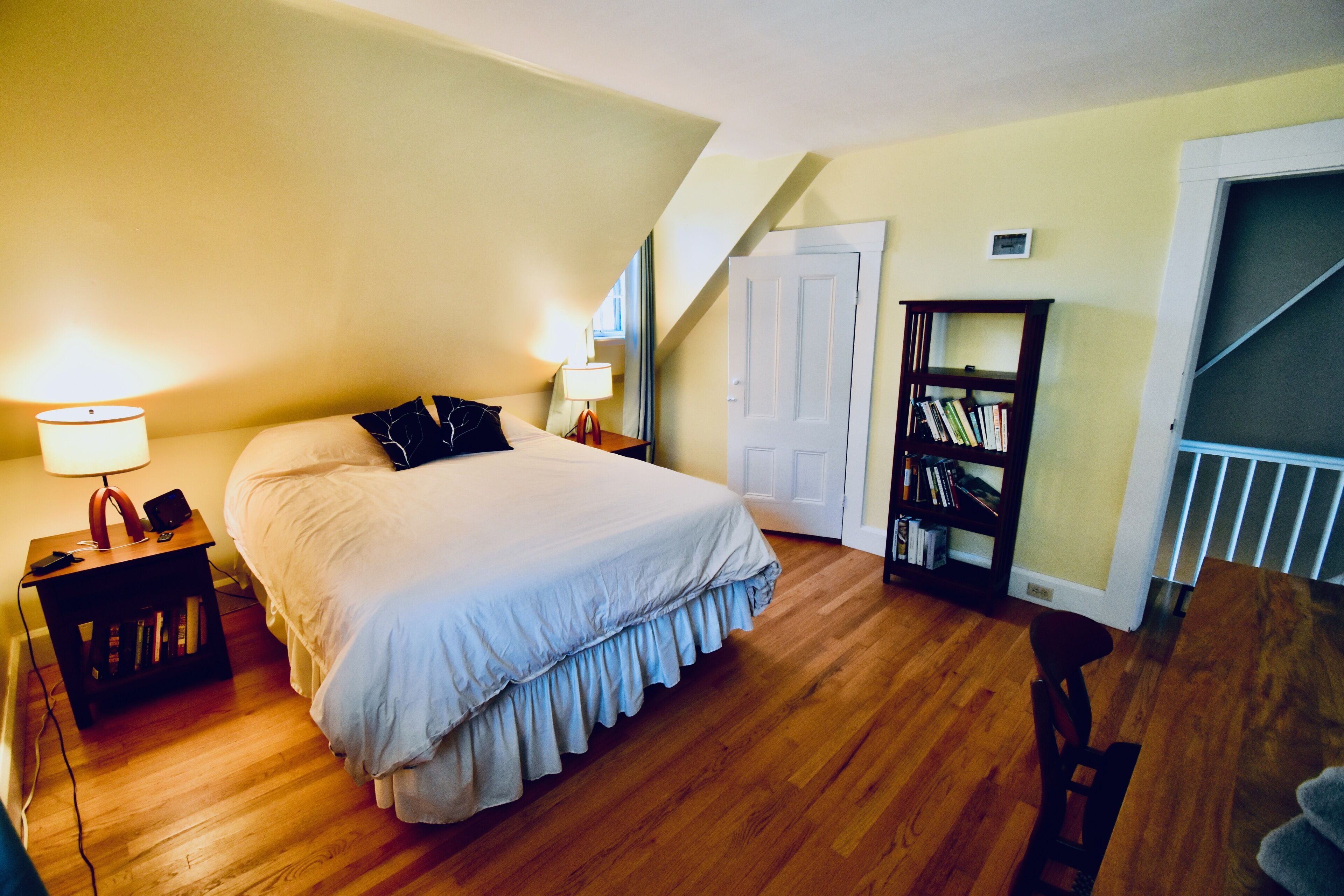 Une chambre à coucher avec un grand lit, une table de chevet avec une lampe, une bibliothèque et une porte donnant sur un escalier.