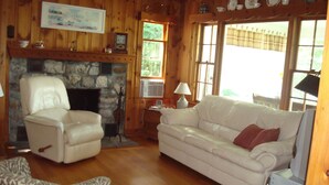 Una sala de estar acogedora con chimenea de piedra, un sillón de cuero blanco, un sofá blanco con un cojín rojo y un reposapiés estampado.