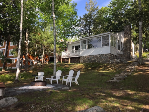 A cabin with a stone foundation and a white exterior, surrounded by trees. There are white Adirondack chairs and a fire pit in the foreground.