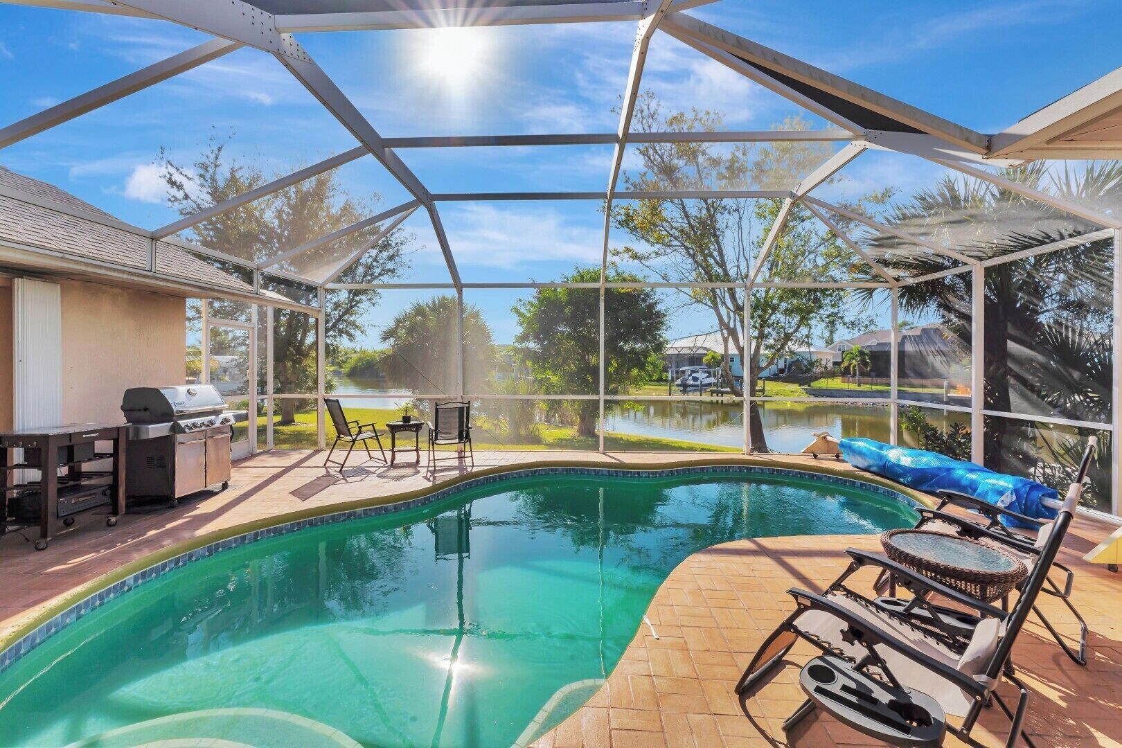 A covered swimming pool with a grill and chairs, surrounded by a view of a lake and trees.