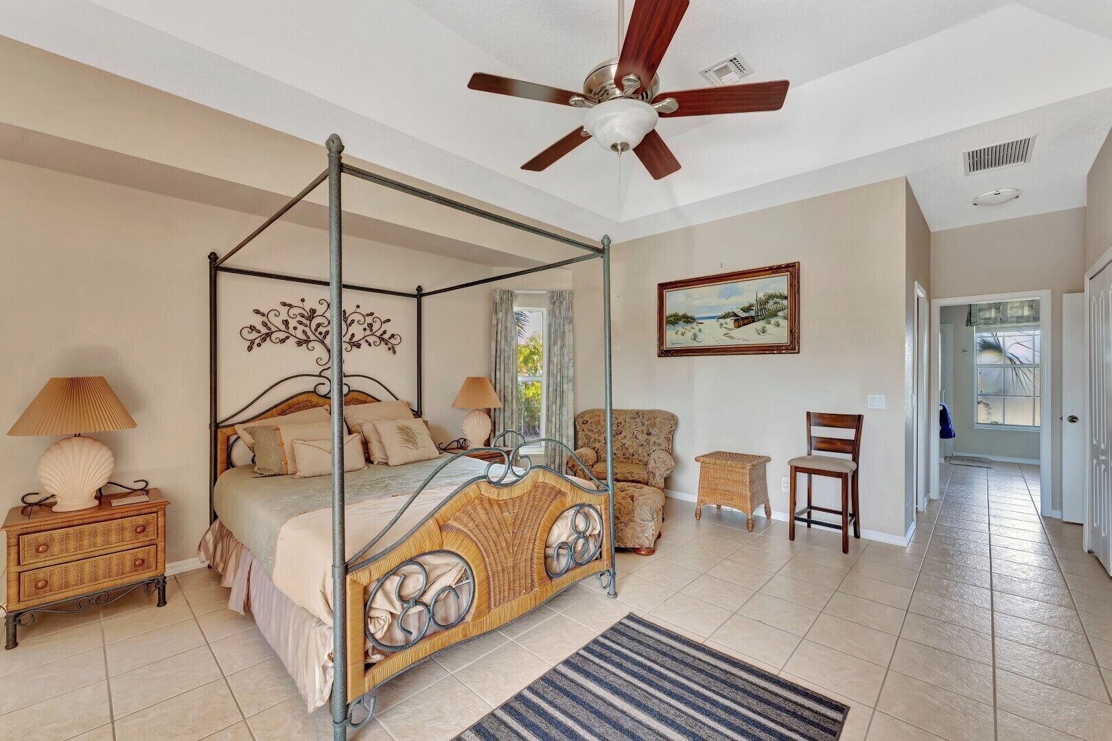 A four-poster bed with a canopy, a ceiling fan, a wicker chair, a small side table, and a painting on the wall.