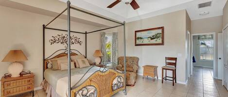 A four-poster bed with a canopy, a ceiling fan, a wicker chair, a small side table, and a painting on the wall.