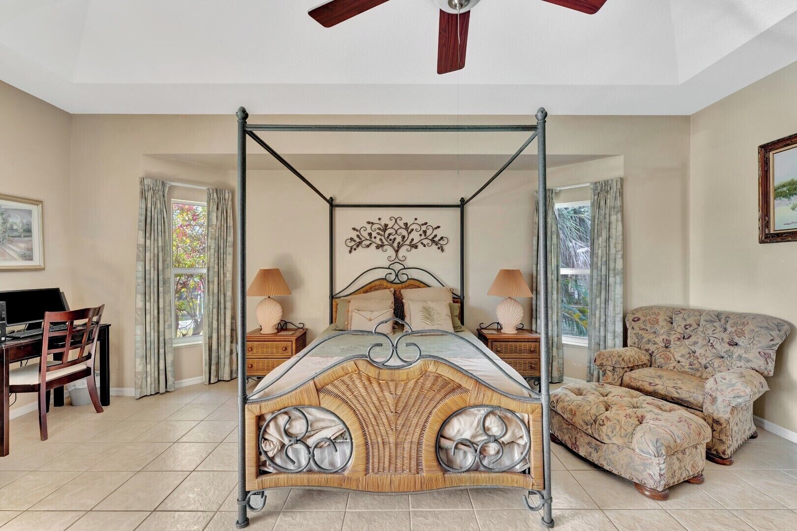 A four-poster bed with a decorative headboard, two bedside tables with lamps, a floral-patterned sofa, and a desk with a chair.