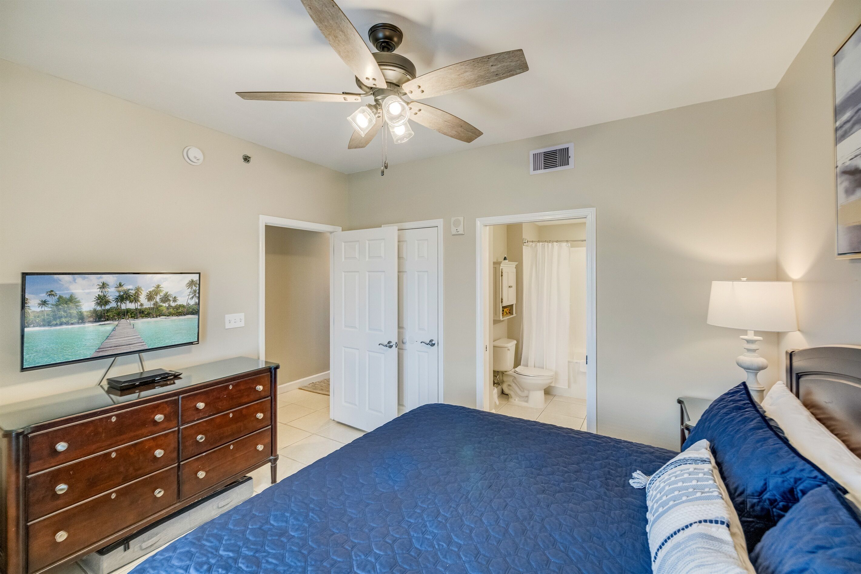 A bedroom with a bed, a ceiling fan, a TV on a dresser, and a bathroom visible through an open door.