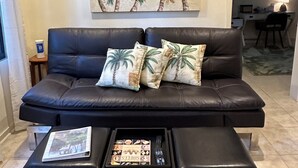 Un salon avec un canapé en cuir noir, une table basse sur laquelle reposent un magazine et une télécommande, un tableau représentant des palmiers accroché au mur, et un bureau avec une lampe.