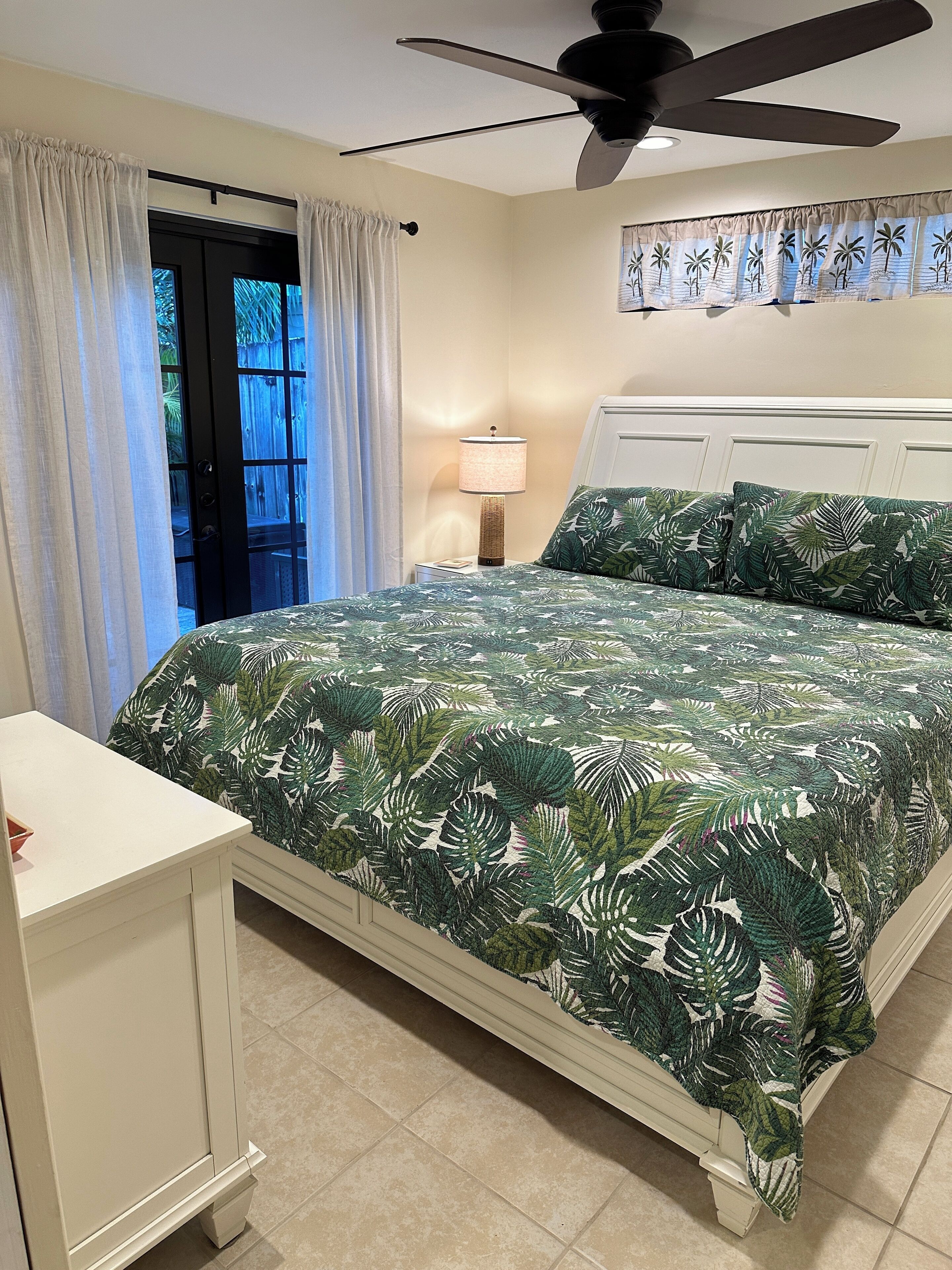 Une chambre à coucher avec un lit, une table de chevet, un ventilateur de plafond et une fenêtre avec des rideaux.