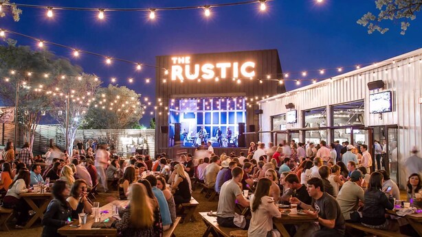 An outdoor dining area with tables and chairs, a stage with a band performing, and a building with 'The Rustic' sign. - Katy Trail Cosy and Elegant (Dallas)