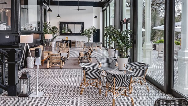Ruime binnenruimte met hoge plafonds, grote ramen en een gevarieerde zithoek, waaronder rieten stoelen en tafels.