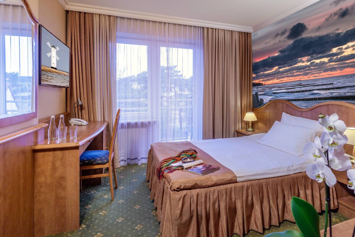 Une chambre d’hôtel comprenant un lit, un bureau, une chaise, une télévision et un tableau représentant un coucher de soleil sur l’océan.