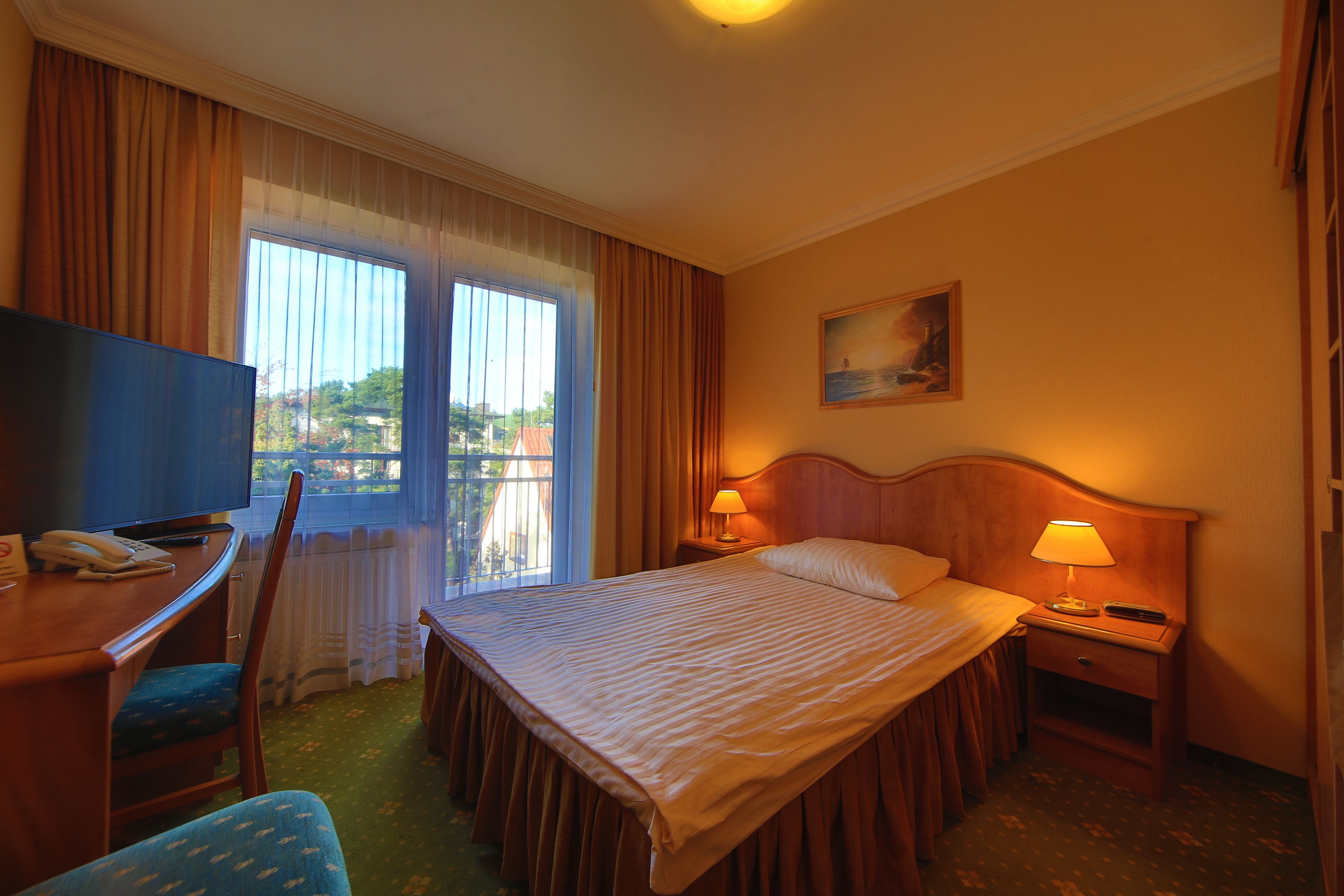 Habitación de hotel con cama, escritorio con televisor, silla, mesita de noche con lámpara y un cuadro en la pared.