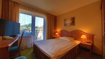Habitación de hotel con cama, escritorio con televisor, silla, mesita de noche con lámpara y un cuadro en la pared.