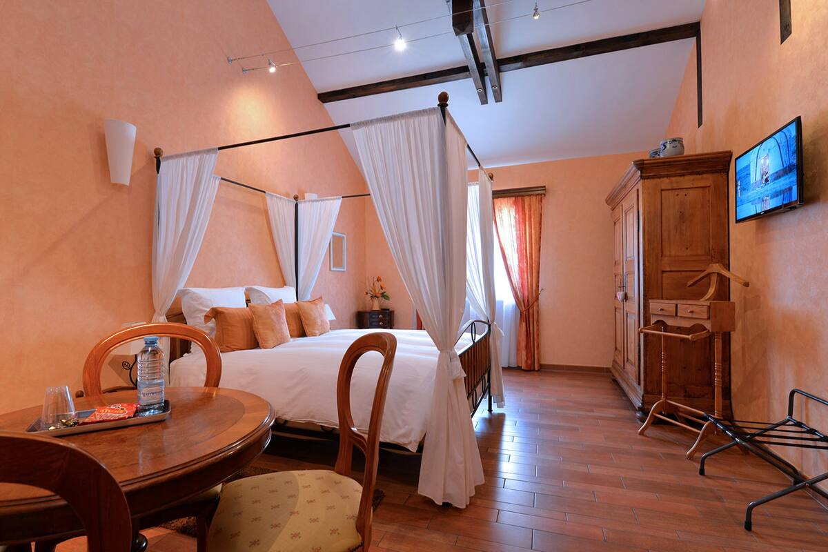 A hotel room with a bed, a wooden chair, a table with a bottle and glass, and a television on the wall.