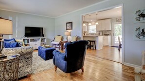 A living room with a sofa, ottoman, and a television. There is a kitchen visible through an open doorway. - Beautiful, Charming, Historic Riverfront and Wrightsville Beach! (Wilmington)