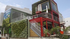 Un edificio moderno con fachada de jardín vertical verde, balcón rojo y blanco, y una escalera que conduce a la entrada.