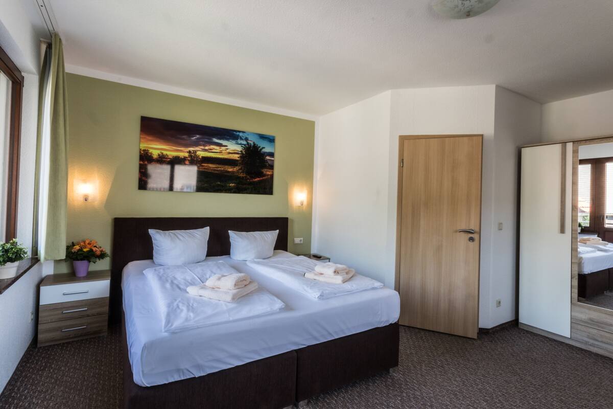 A hotel room with a large bed, a nightstand with a lamp, a painting on the wall, and a wardrobe.