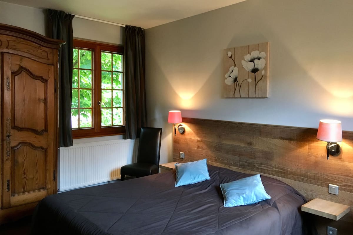Een slaapkamer met een groot bed, twee bedlampen, een houten hoofdbord, een stoel, een raam met gordijnen en een radiator.