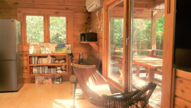 A wooden cabin interior with a kitchen, a bookshelf, a hammock, and a sliding glass door leading to an outdoor deck. - YOISAMA Sunrise Beach House (Ishigaki)