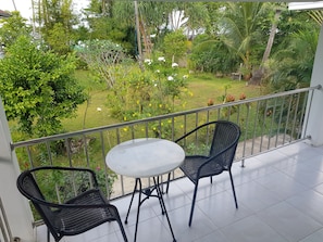 A balcony with two black wicker chairs and a small round table, overlooking a lush garden with various plants and flowers. - Mook Ing Lay (Ko Mook)
