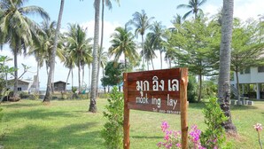 A wooden sign with text in a non-Latin script, surrounded by palm trees and a grassy area. - Mook Ing Lay (Ko Mook)
