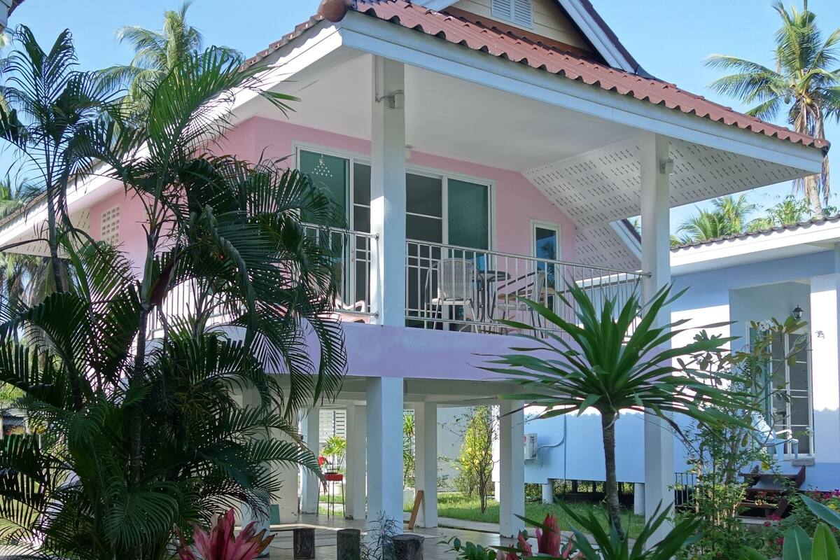 A two-story house with a balcony, surrounded by tropical plants and a clear blue sky.