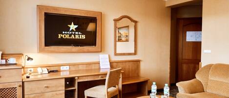 A hotel room with a wooden desk, a chair, a sofa, a TV mounted on the wall, and a small table with water bottles.