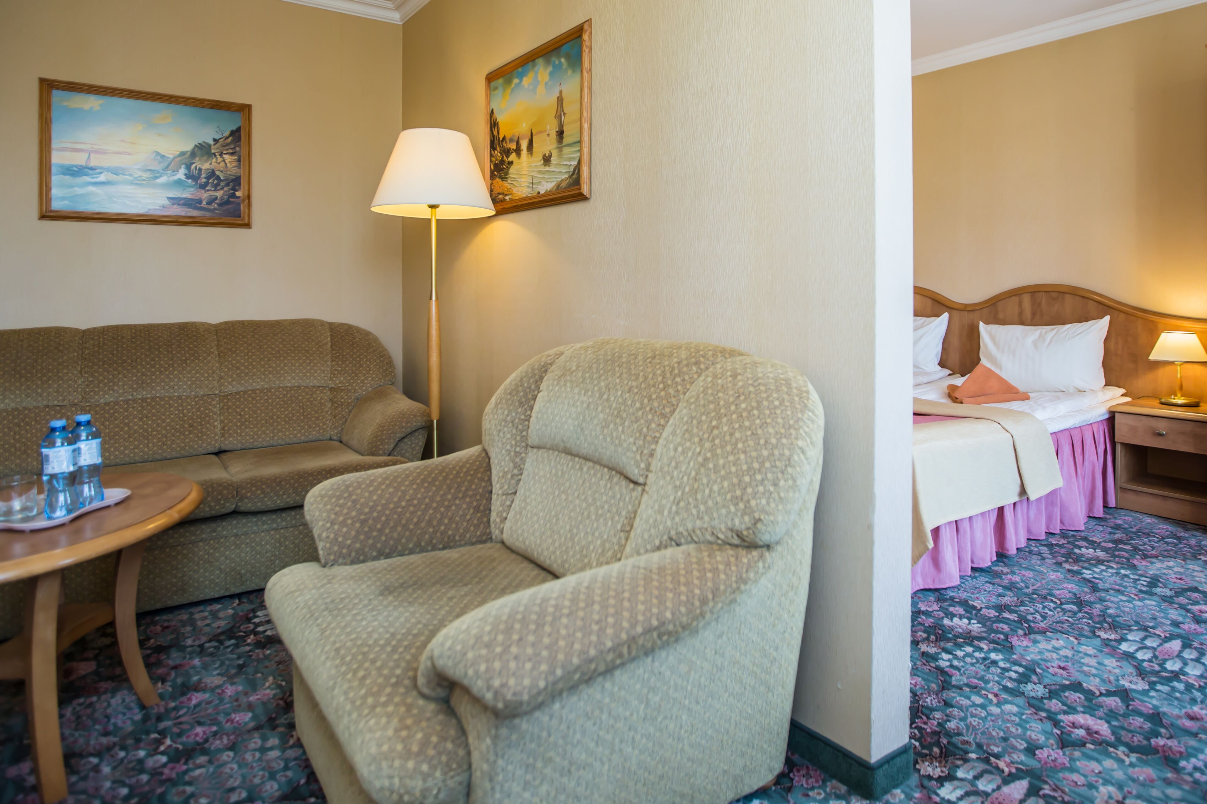Habitación de hotel con sofá, sillón y cama con falda rosa. Hay dos cuadros en la pared y una lámpara en la mesita de noche.