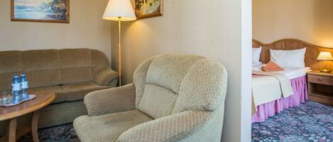 A hotel room with a sofa, armchair, and a bed with a pink skirt. There are two paintings on the wall and a lamp on the side table.