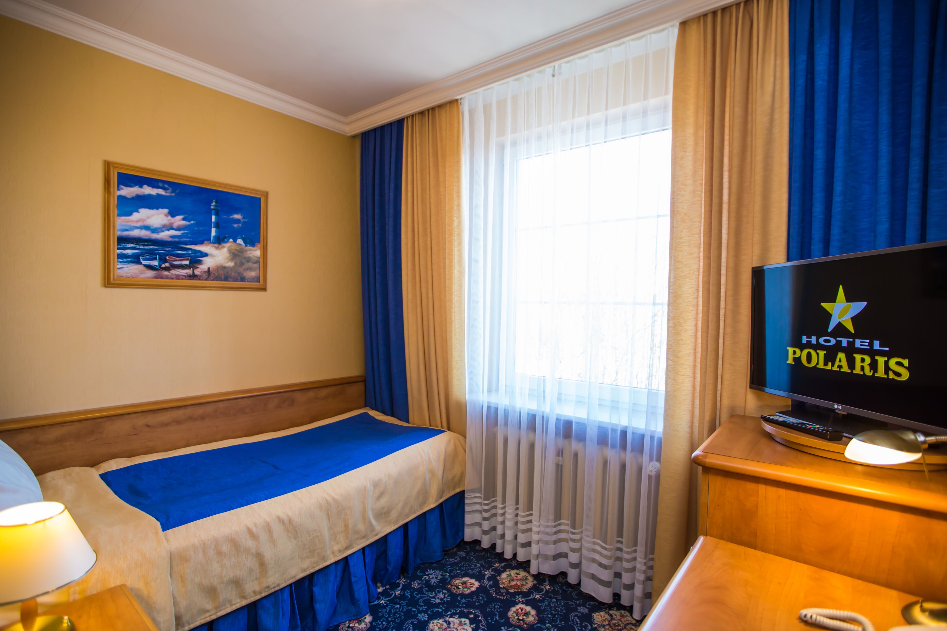 A hotel room with a bed, a television displaying the hotel name, a painting on the wall, and a window with curtains.