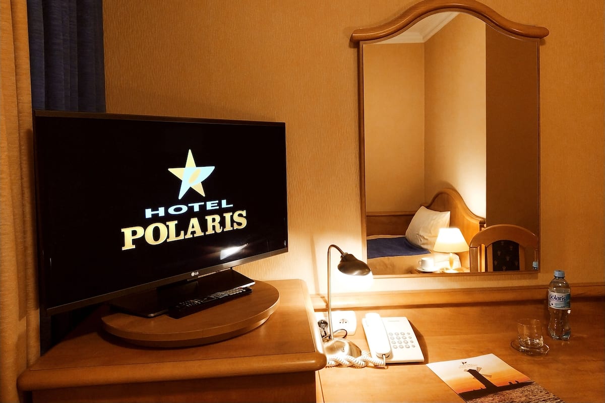Habitación de hotel con una televisión mostrando el logo del Hotel Polaris, una lámpara de noche, un teléfono, una botella de agua y un espejo que refleja el interior de la habitación.