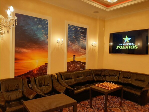 A hotel room with a large sofa, a chess table, and two wall-mounted screens displaying the hotel name.