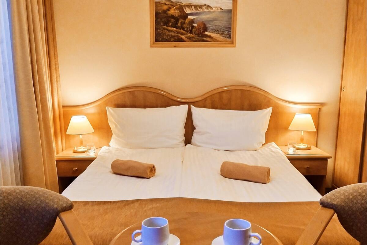 Habitación de hotel con una cama doble, dos mesitas de noche con lámparas, una mesita redonda pequeña con dos tazas y dos sillas estampadas.