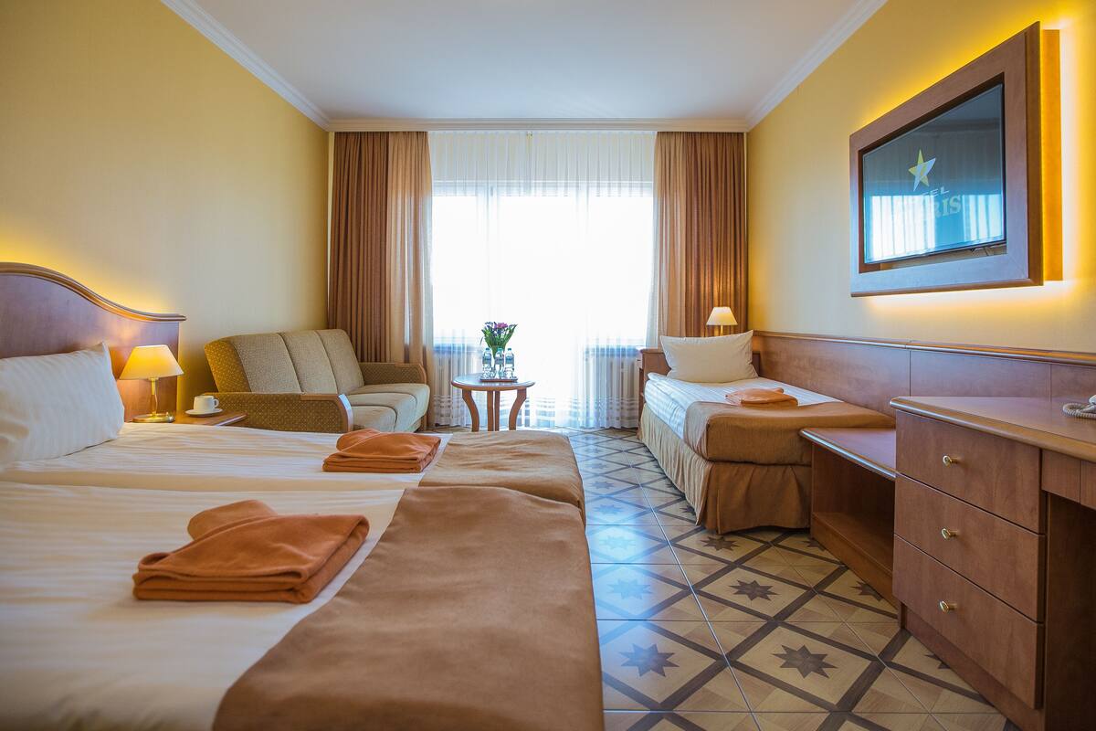 Habitación de hotel con dos camas, un sofá, una mesita con un florero y un televisor en la pared.