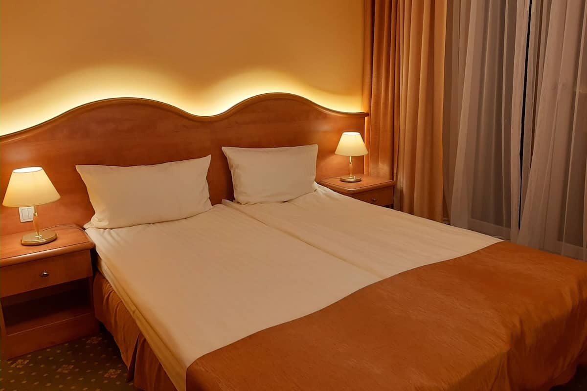 Habitación de hotel con una cama grande, dos mesitas de noche con lámparas, un cabecero de madera y un ventanal con cortinas.