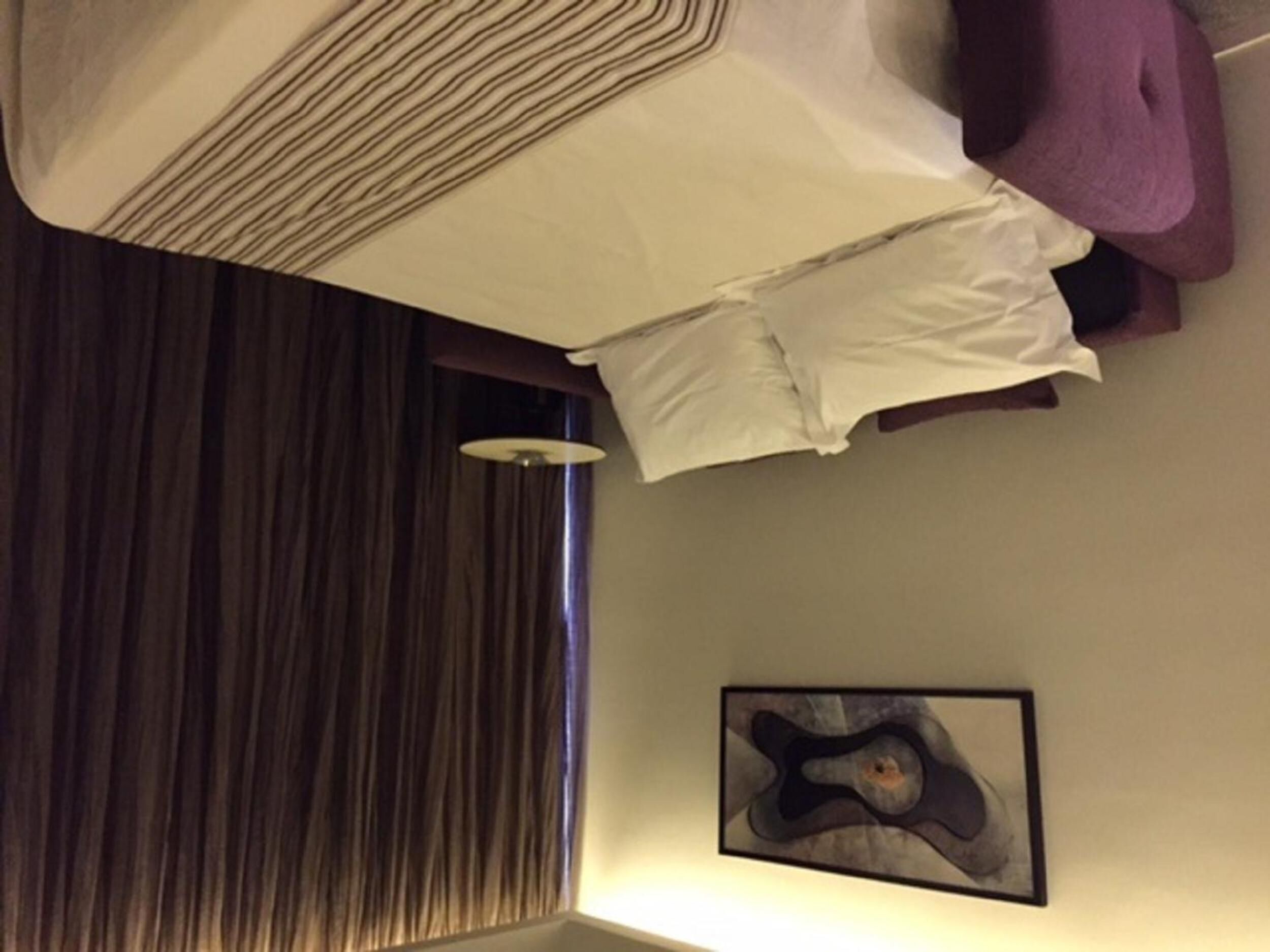 Quarto de hotel com cama, uma cadeira roxa, uma obra de arte na parede e janela com cortinas.
