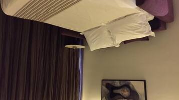 Quarto de hotel com cama, uma cadeira roxa, uma obra de arte na parede e janela com cortinas.
