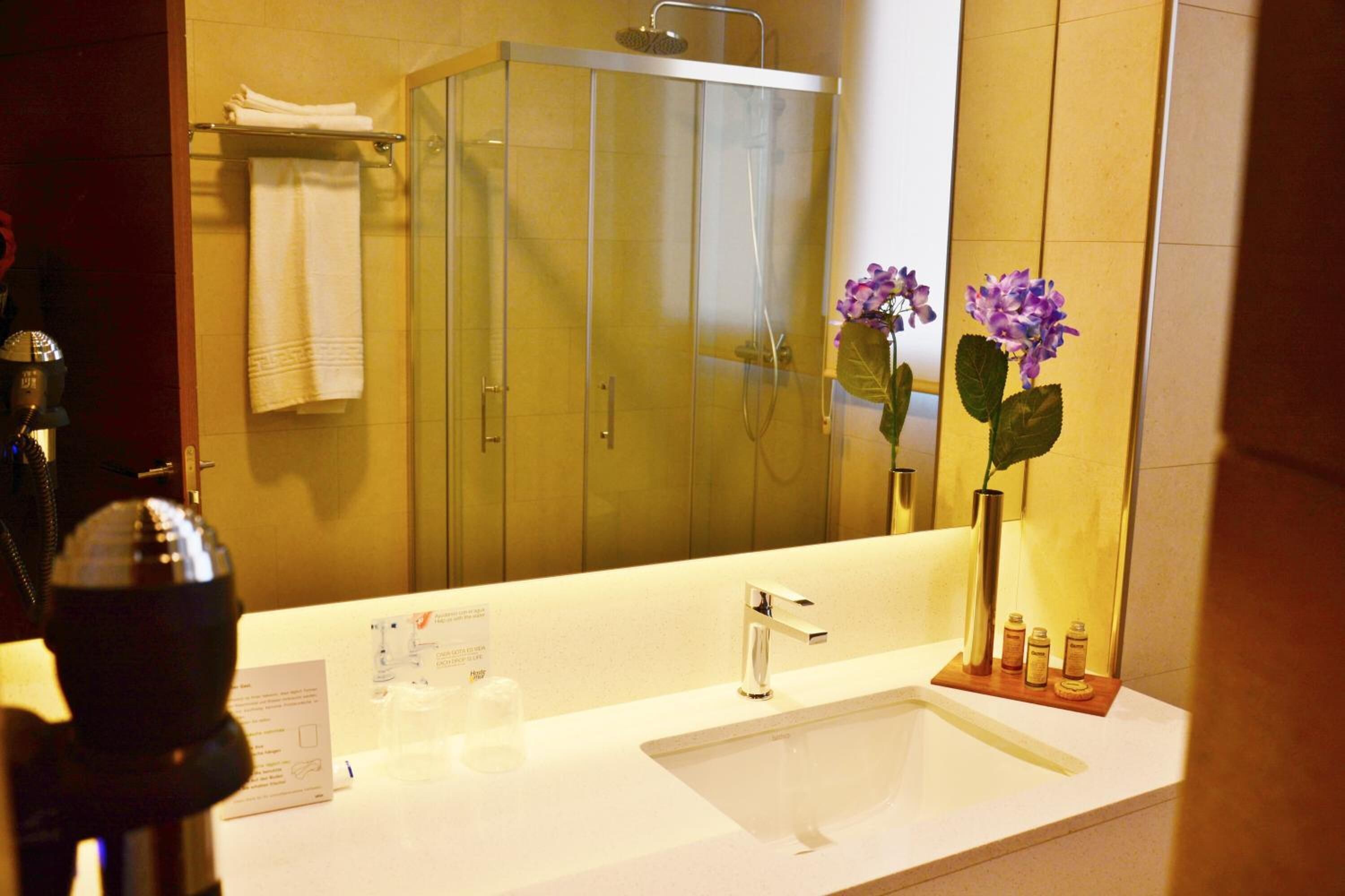 Un baño con un espejo grande, un lavabo, una ducha y un florero con flores.
