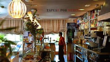 A bar area with a counter displaying food and drinks, a person standing behind the counter, and shelves stocked with bottles and glasses.