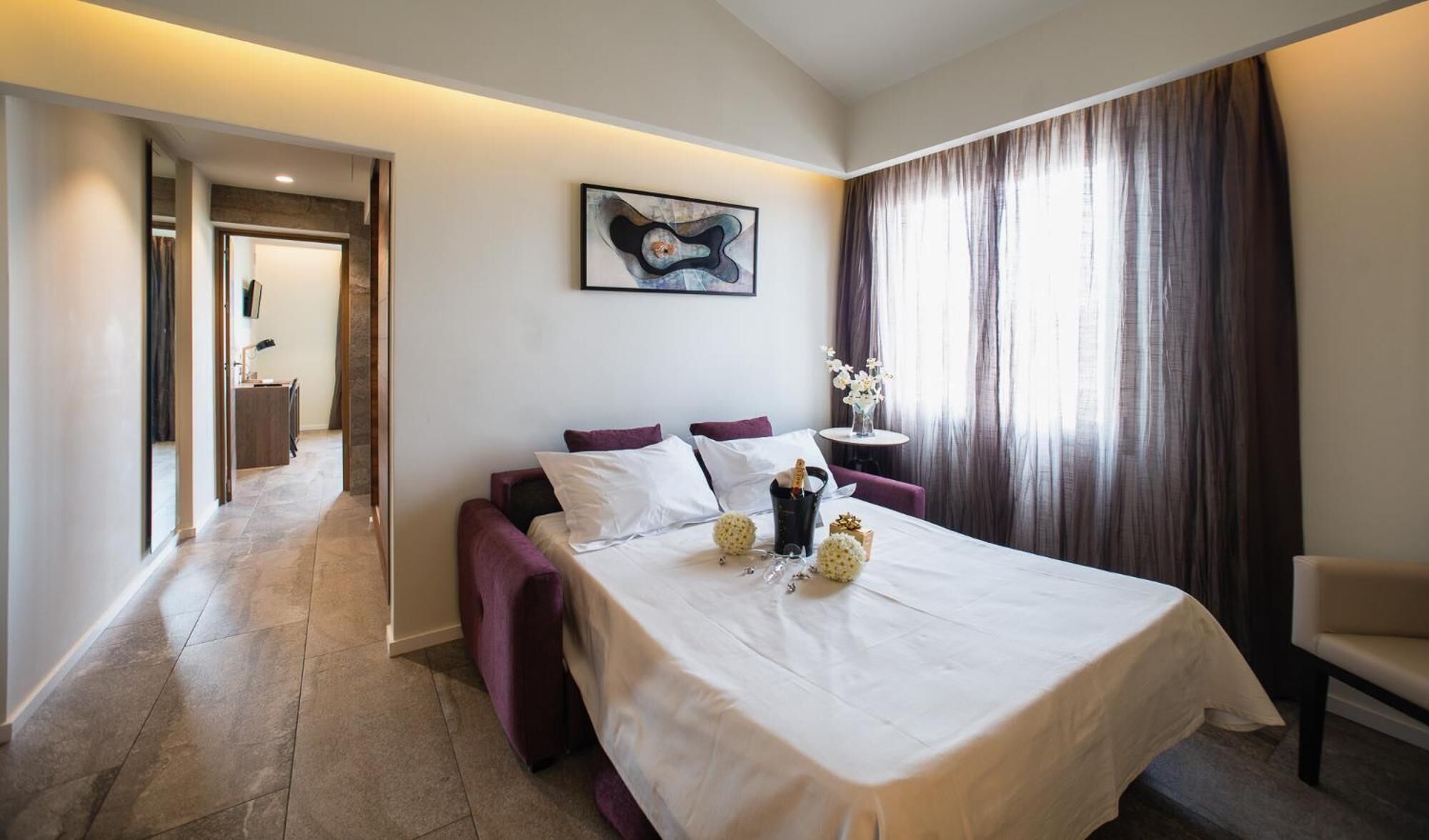 A hotel room with a bed, a small table with a vase of flowers, and a painting on the wall.