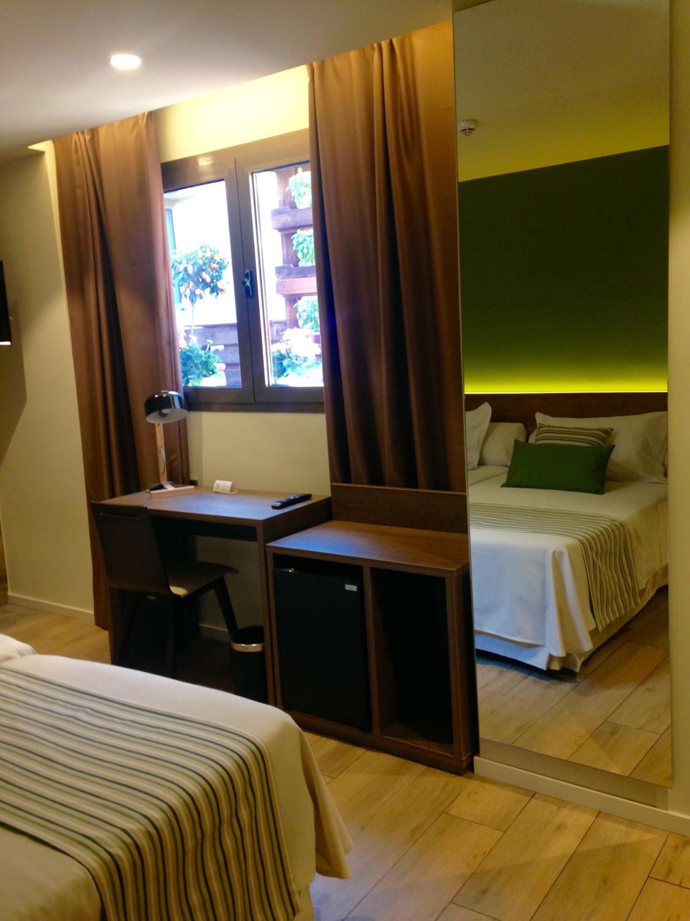 A bedroom with a bed, a desk, and a chair. There is a window with curtains, a lamp, and a view of greenery outside.