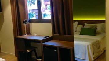 A bedroom with a bed, a desk, and a chair. There is a window with curtains, a lamp, and a view of greenery outside.