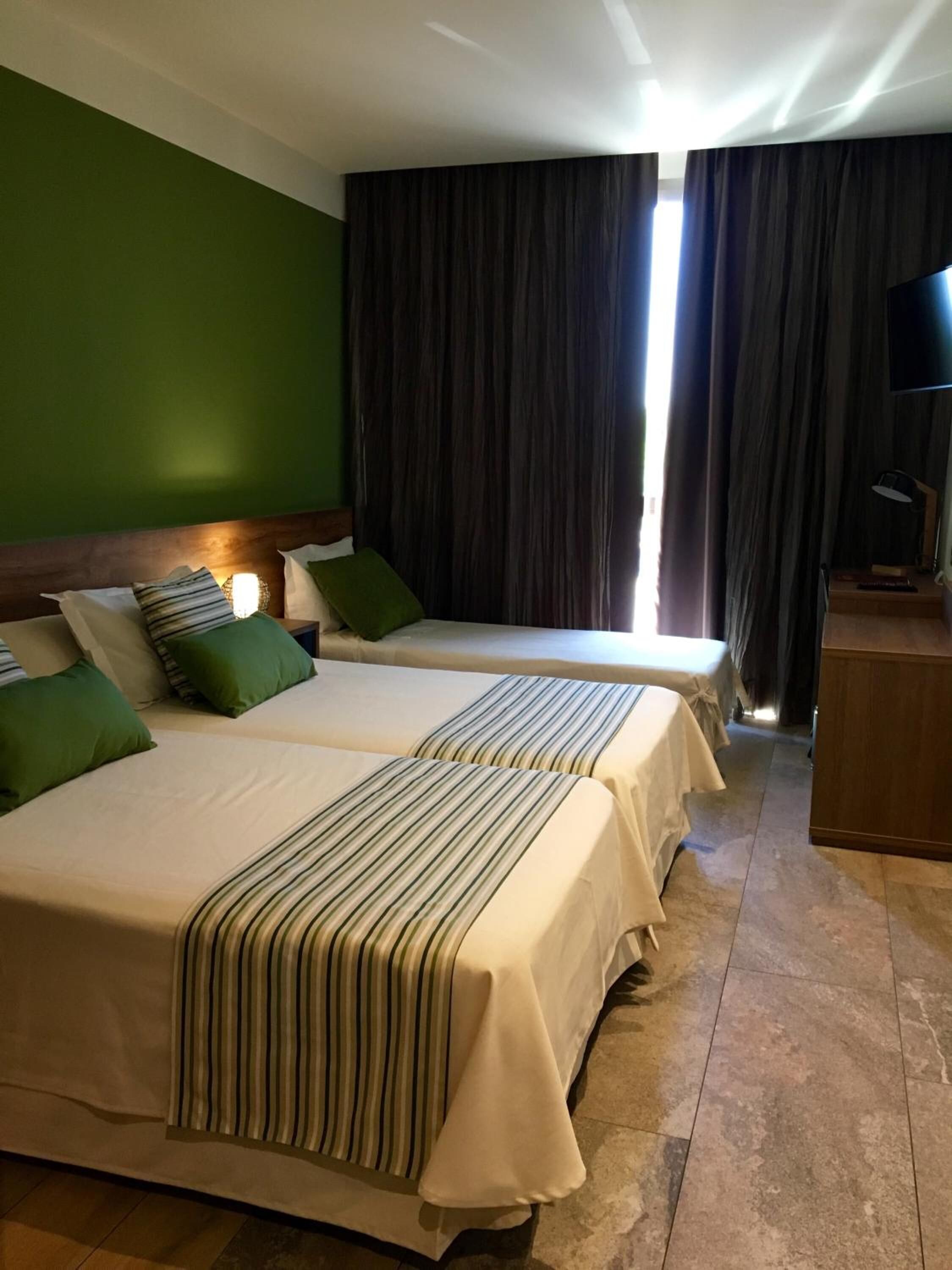 Habitación de hotel con dos camas, paredes verdes, un ventanal grande con cortinas, un televisor de pantalla plana y una mesita de noche de madera.