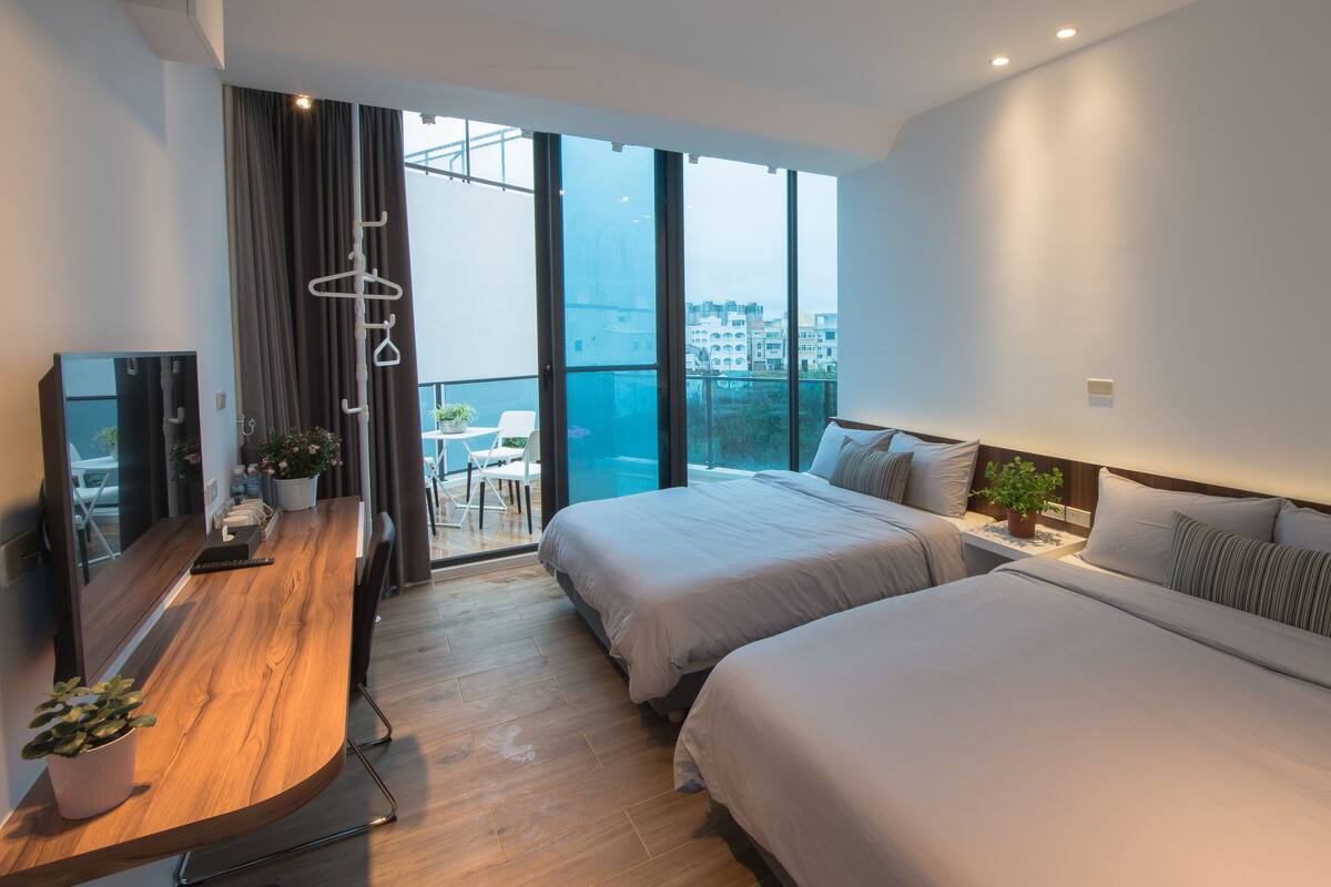 A modern hotel room with two beds, a wooden desk, a TV, and a balcony with a city view.